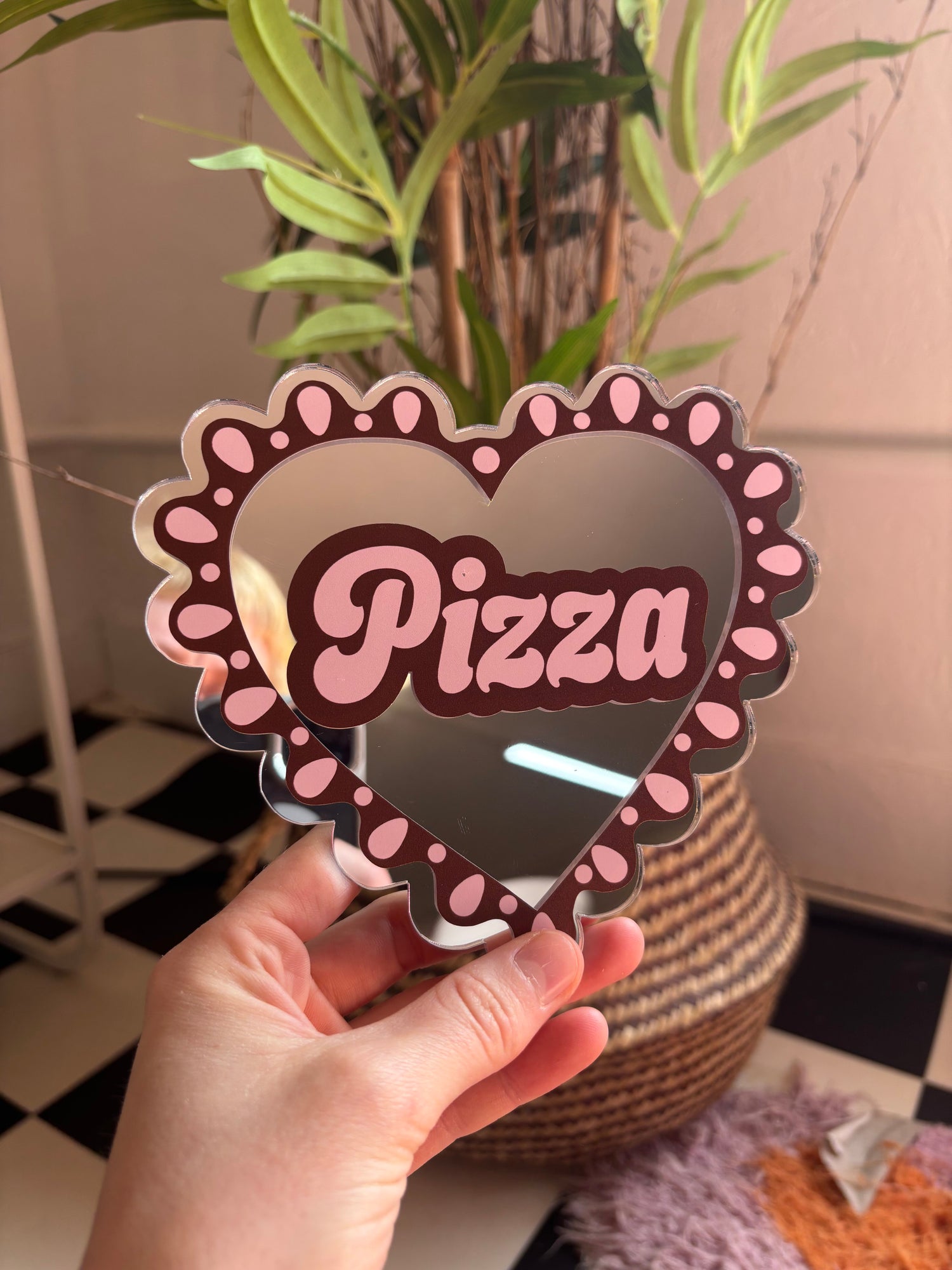A hand holding a heart-shaped sign with the word "Pizza" written on it, in front of a potted plant.