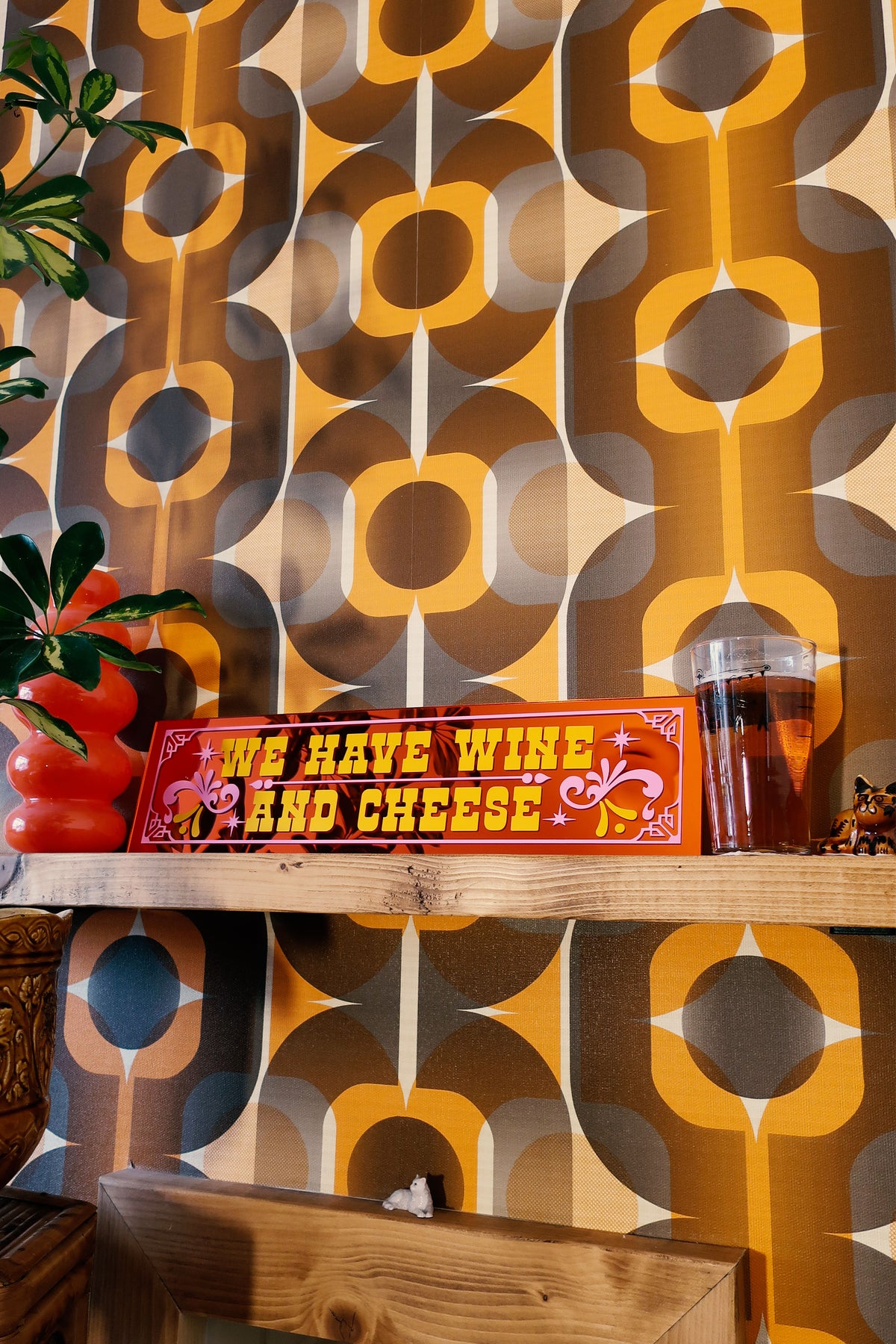 a shelf with a sign that reads "We have wins and cheese", a glass of beer, and a plant. The background has a patterned wallpaper with a repeating design of circles and lines in various shades of orange and brown.