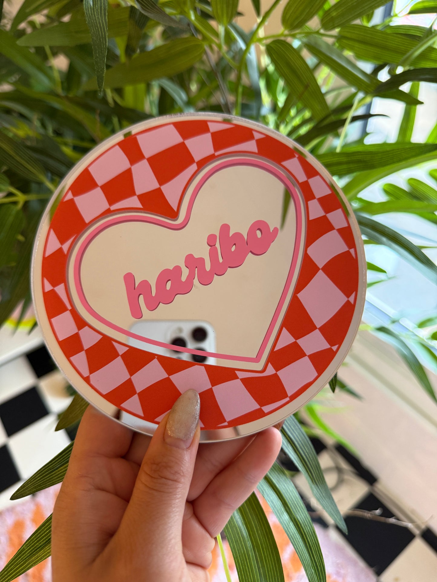 A hand holding a heart-shaped mirror with the word "Love" written on it, against a backdrop of green leaves and a checkered pattern.