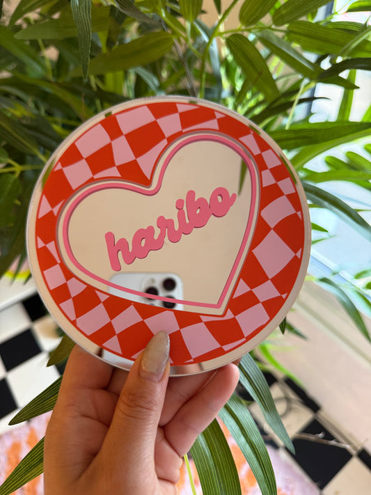 A hand holding a heart-shaped mirror with the word "Love" written on it, against a backdrop of green leaves and a checkered pattern.