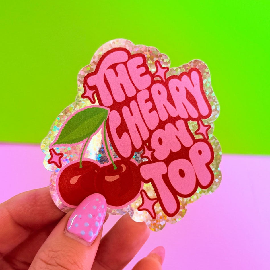 Sticker with 'The Cherry on Top' text held by a hand against a green and pink gradient background