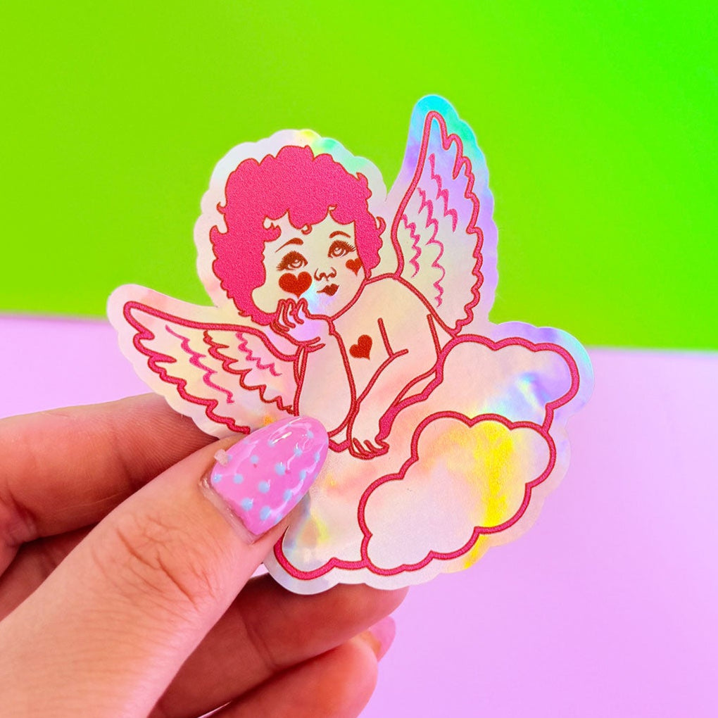 Hand holding a colorful sticker of a cartoon character with wings against a gradient green and pink background.