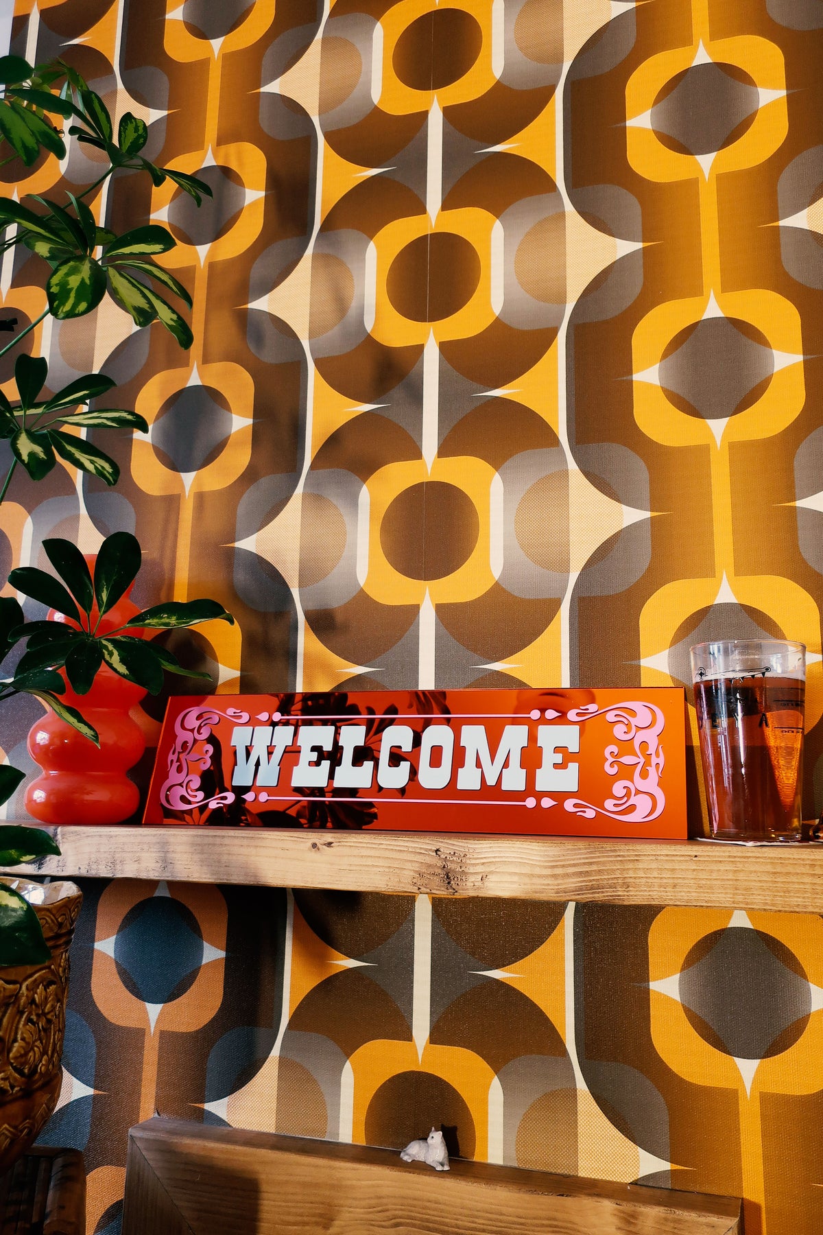 a wooden shelf with a red sign that says "WELCOME", a glass of what appears to be a beverage, and a potted plant. The background has a patterned wallpaper with a repeating design of circles and lines in various shades of orange and brown.