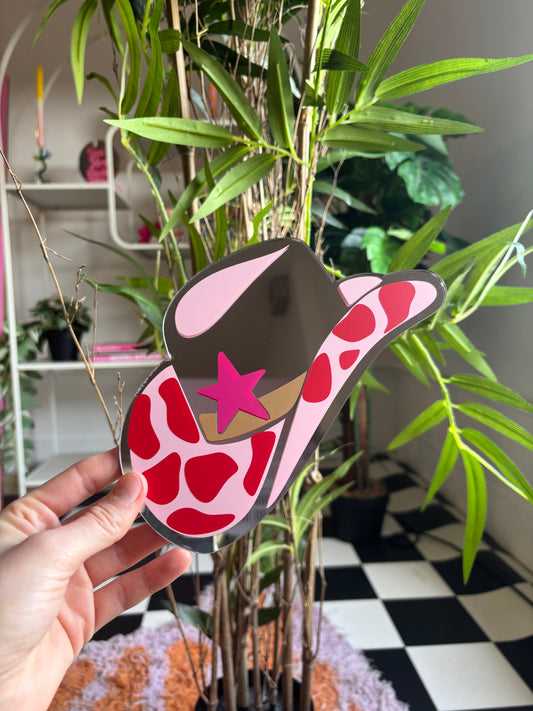 A hand is holding a colorful, cartoonish cowboy hat with a pink star on it, in front of a plant with green leaves and a checkered floor.