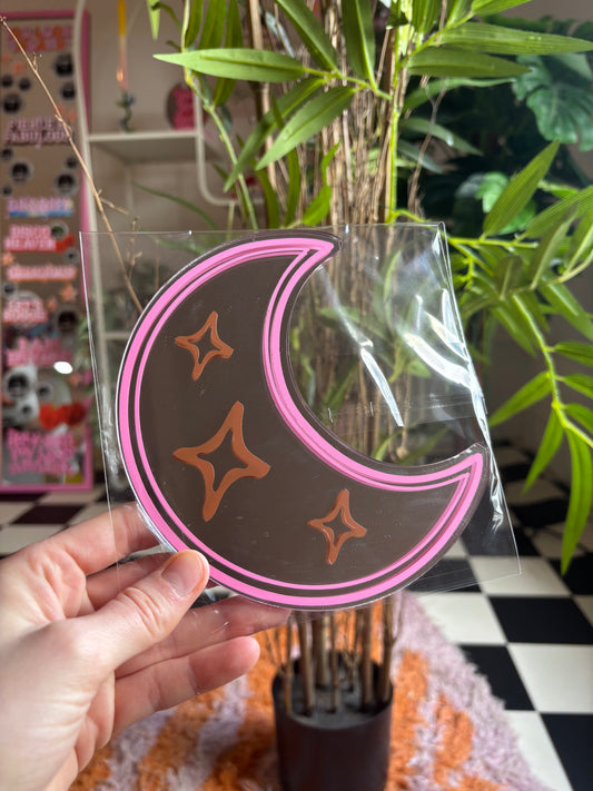 A hand is holding a crescent moon-shaped object with three stars on it, which is placed in front of a potted plant.
