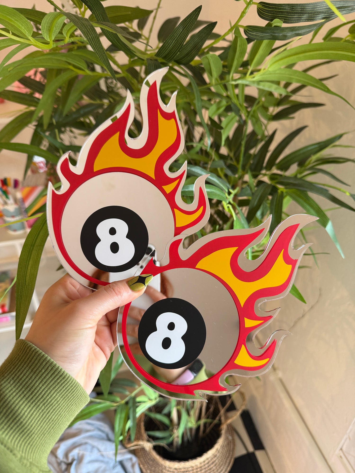 A hand holding a pair of colorful, flame-shaped mirrors against a backdrop of greenery.