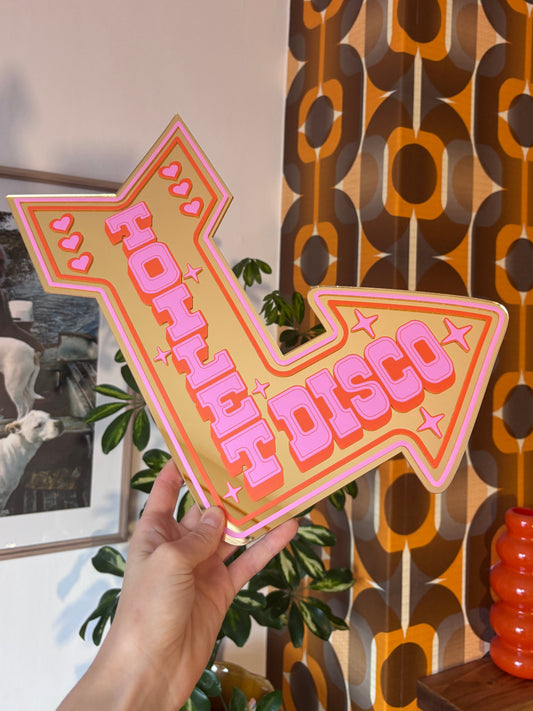 A hand is holding a pink and yellow sign with the word "HAPPY" written on it, which is positioned in front of a wall with a retro-style pattern.