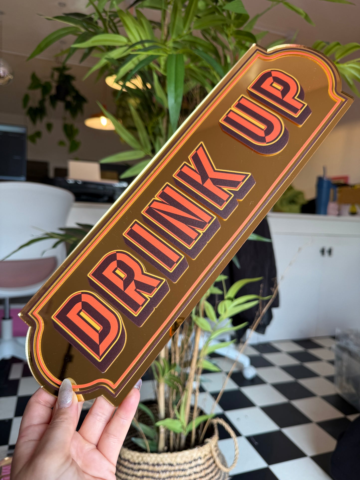 A person is holding a sign that says "DRINK UP" in a room with a checkered floor and plants.