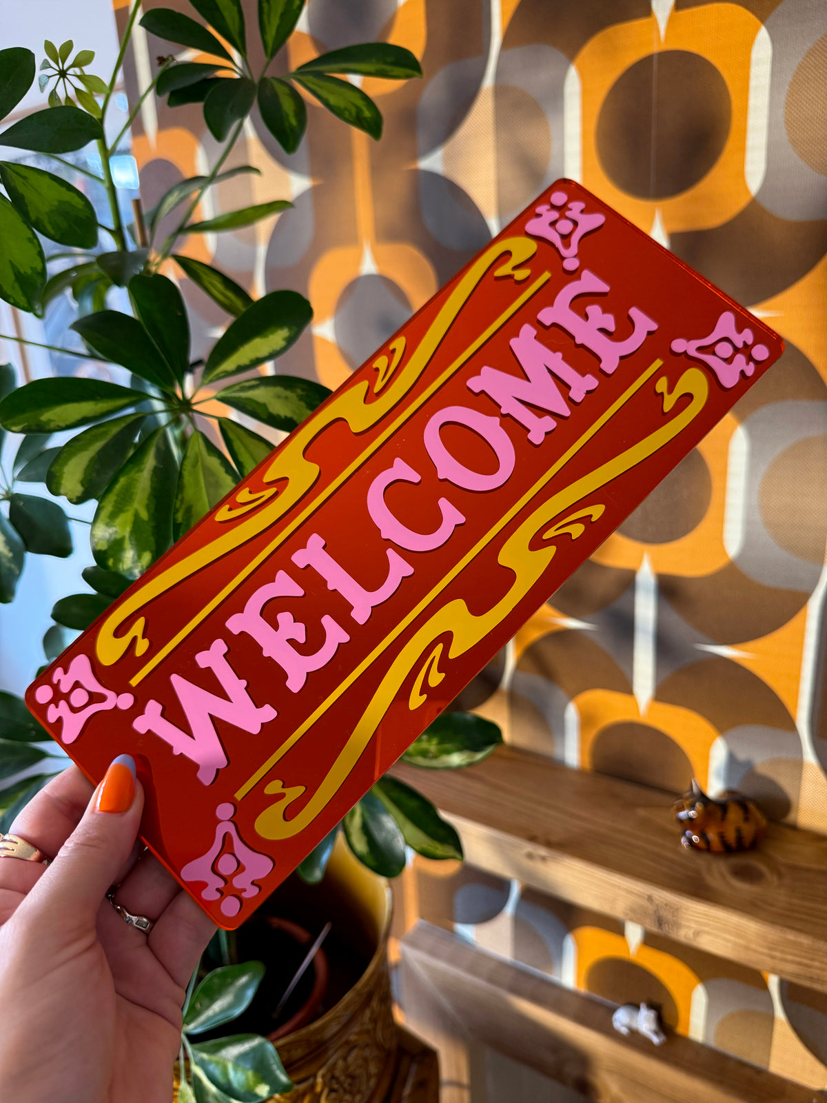 A person is holding a red sign with the word "WELCOME" written on it, in front of a plant and a shelf with various items.