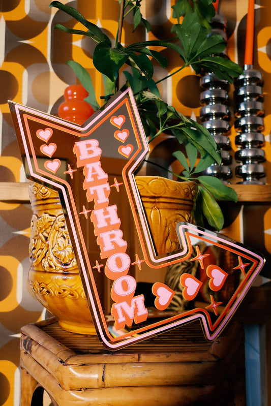 a bathroom sign with a pink and orange design, placed on a wooden stool. The sign has the word "BATHROOM" written on it, and there are heart-shaped decorations surrounding the sign. The background includes a patterned wall and a potted plant.