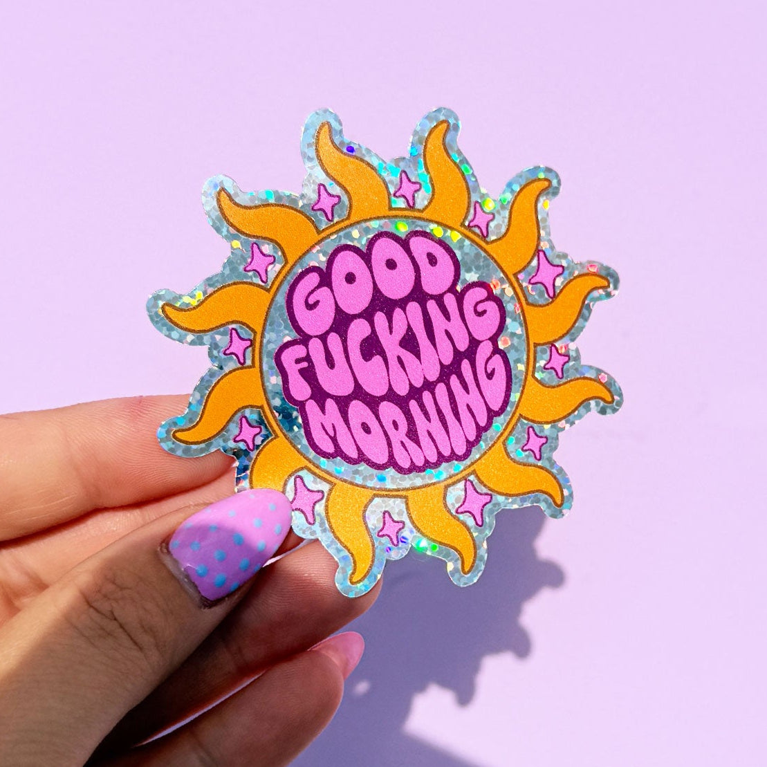 Hand holding a colorful sticker with 'Good Fucking Morning' text against a light purple background
