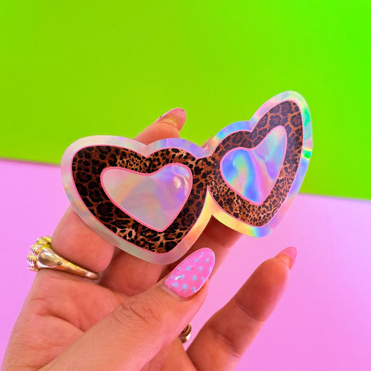 Heart-shaped sunglasses with leopard print details held by a hand against a green and pink gradient background.
