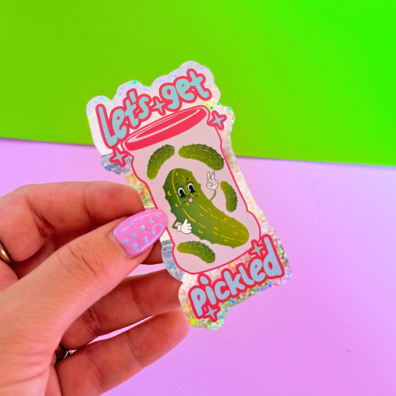 Hand holding a sticker with a pickle design against a colorful background