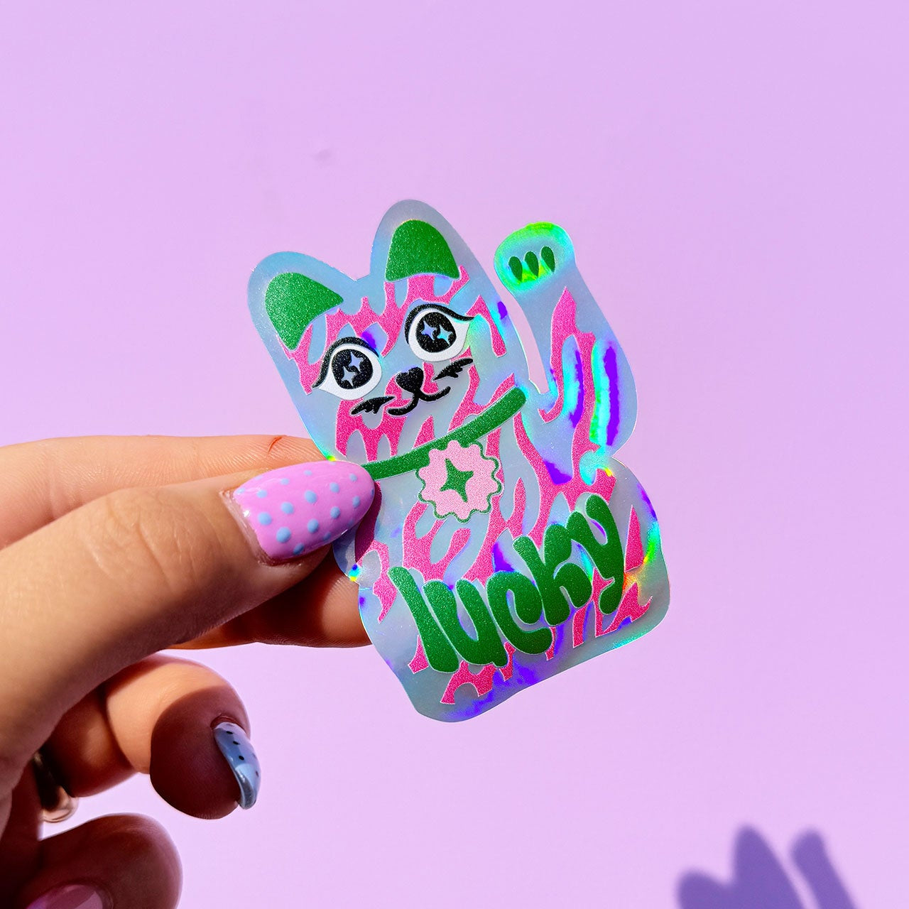 Hand holding a colorful cat-shaped sticker with 'lucky' text on a purple background