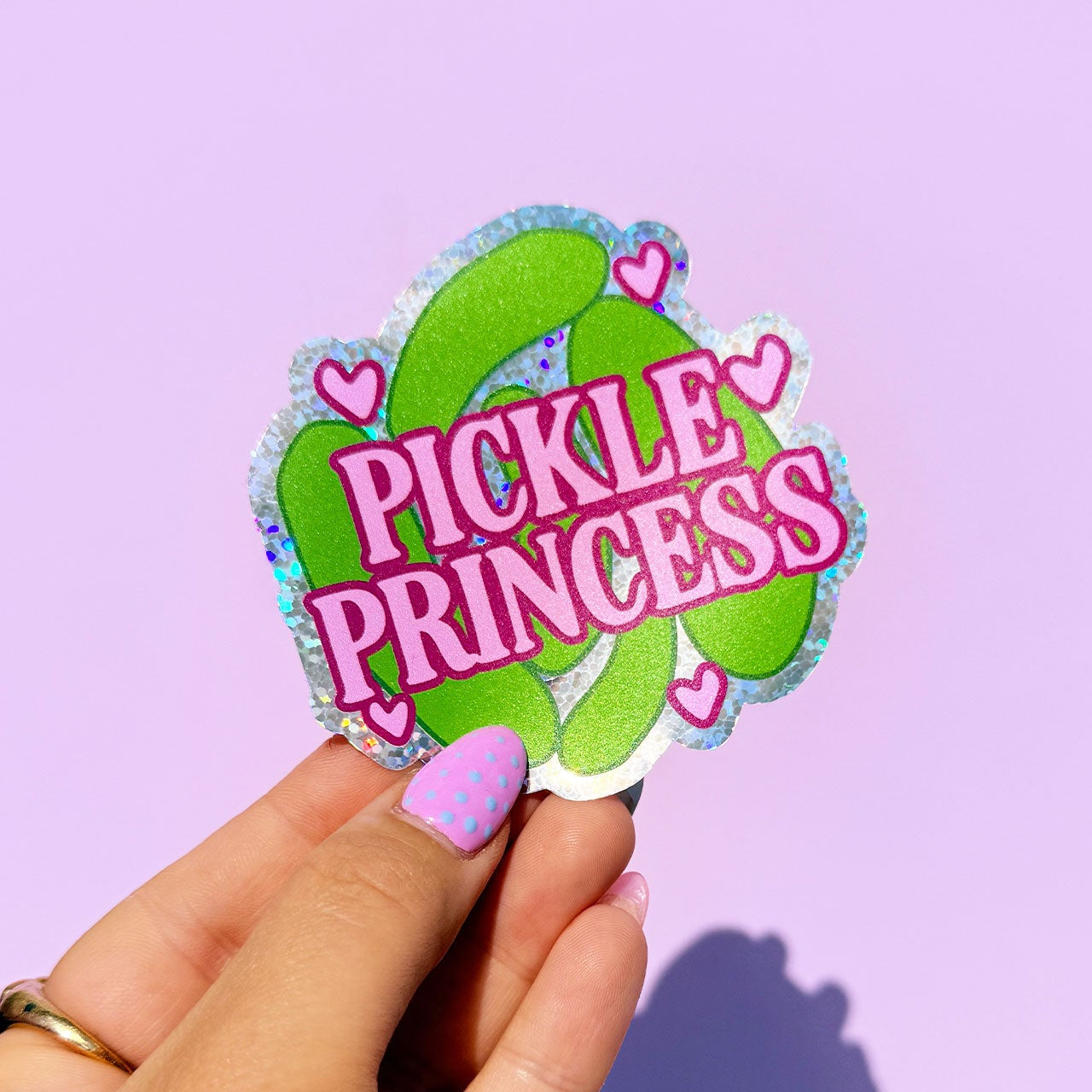 Hand holding a 'Pickle Princess' sticker against a purple background