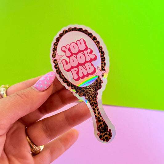 Hand holding a decorative mirror with 'You Look Fab' text against a colorful background